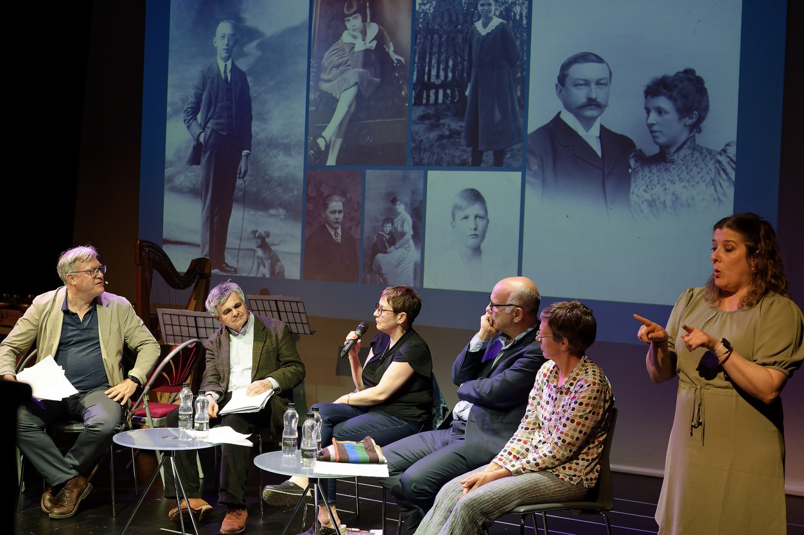 A group of people are seated in front of a screen, one is speaking into a microphone. A woman is standing and signing.