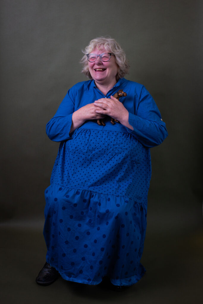 Smiling woman in a blue dress holding a small toy horse against a plain background.