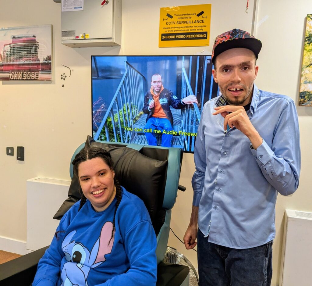 Two people standing indoors singing with a TV screen showing their music video behind them.