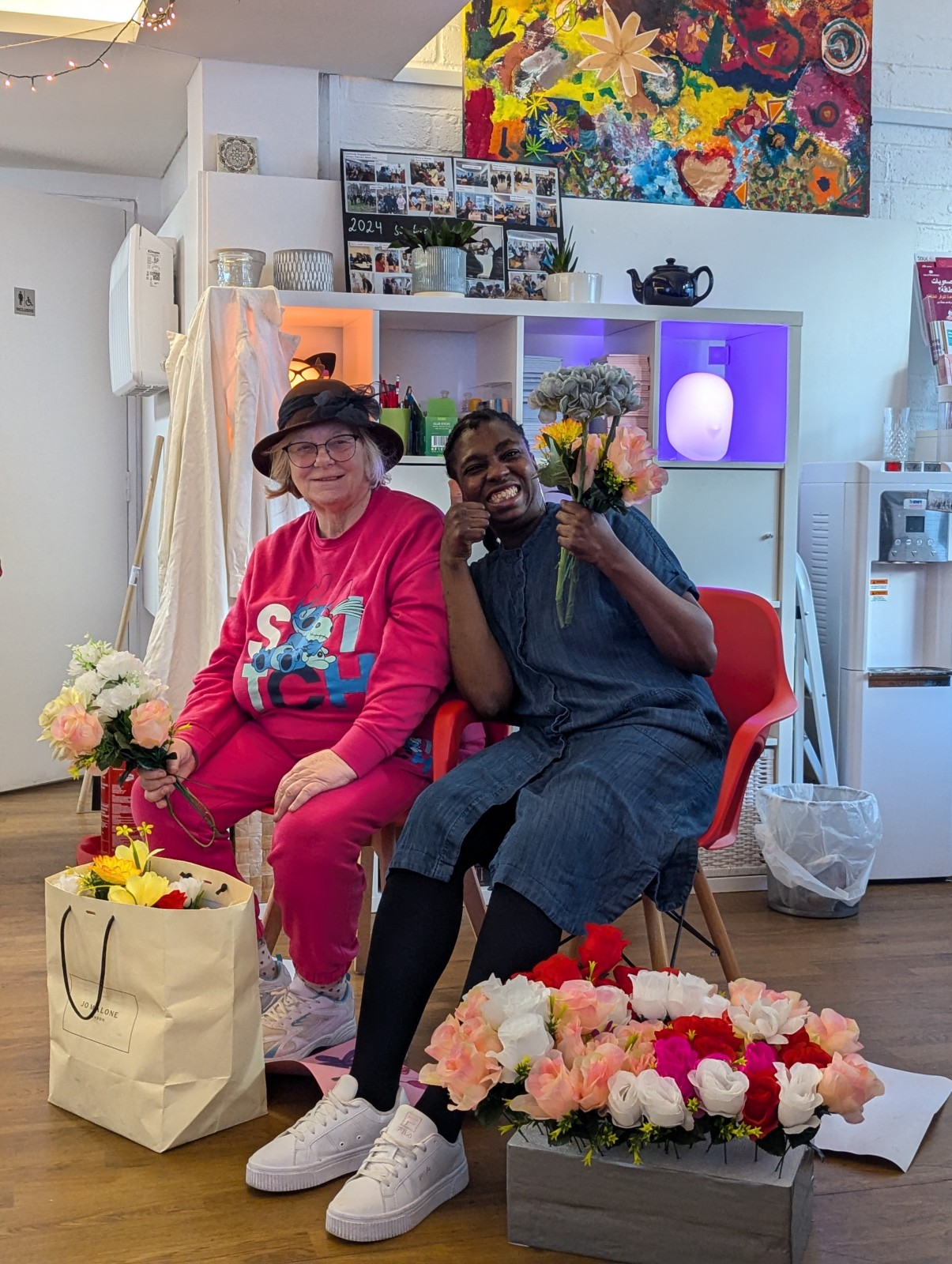 Two people sitting indoors smiling while holding bouquets of flowers.