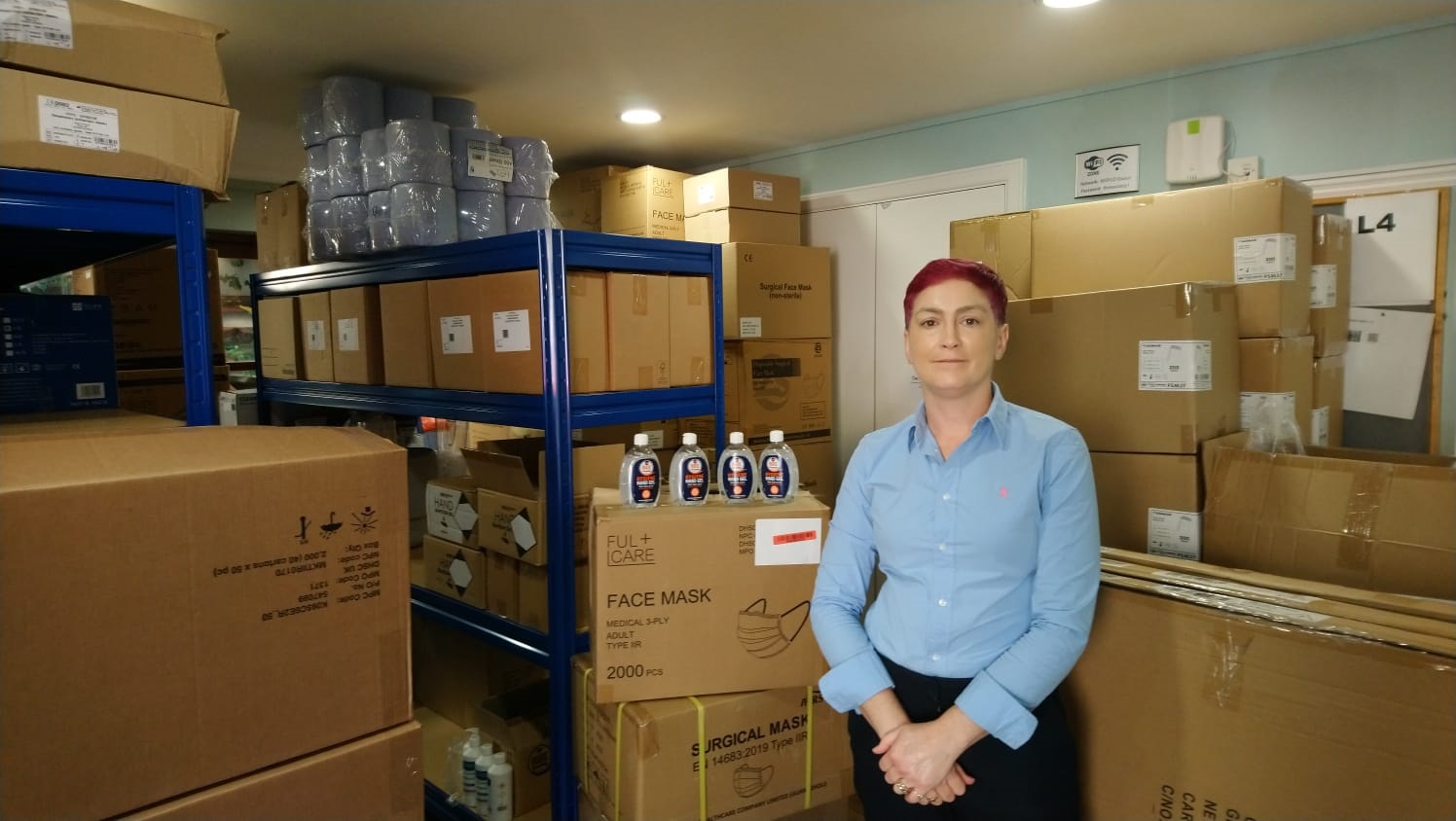 Clare stands in a store cupboard surrounded by boxes full of PPE equipment