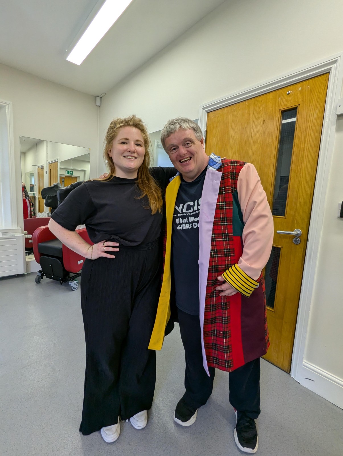 Two people smiling with their arms around each other in a bright room.