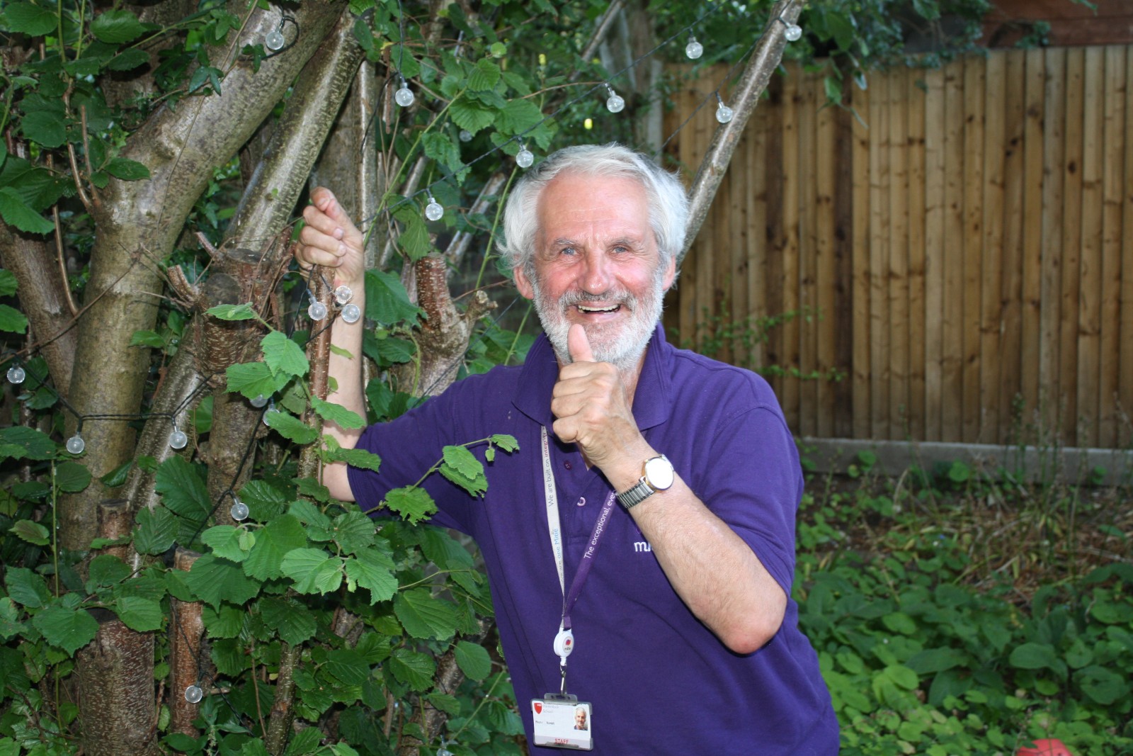 A man is standing in a garden next to a tree, he is smiling and has his thumb up