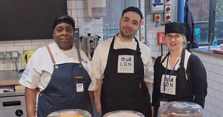 Three kitchen staff standing behind a counter with cakes and pastries on display.