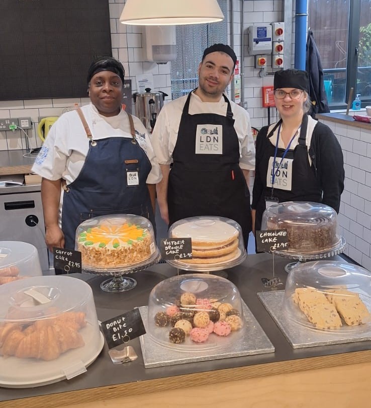 Three kitchen staff standing behind a counter with cakes and pastries on display.