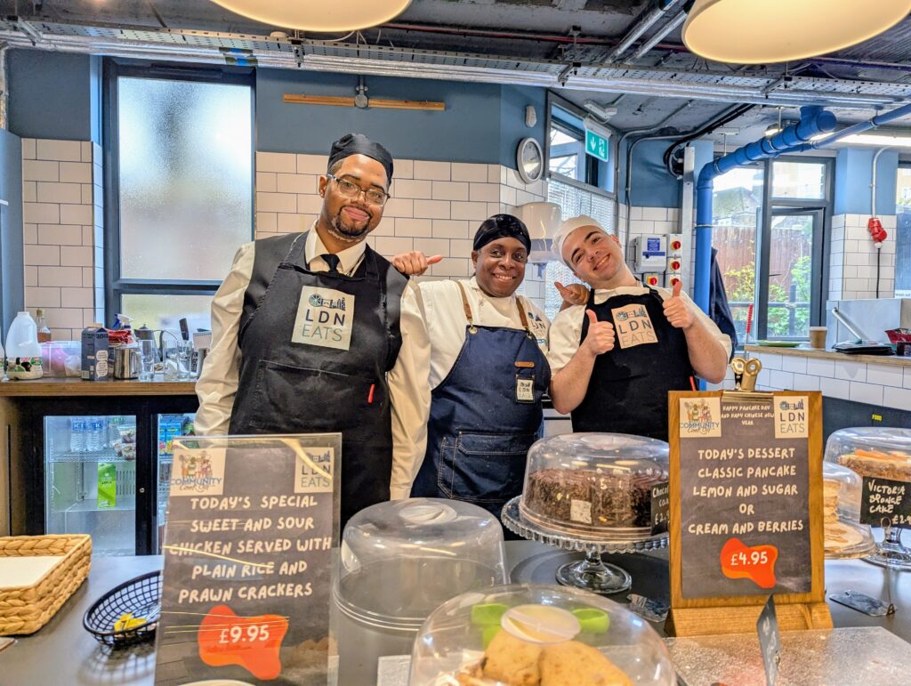Three LDN Eats staff smiling behind a café counter with dessert displays.
