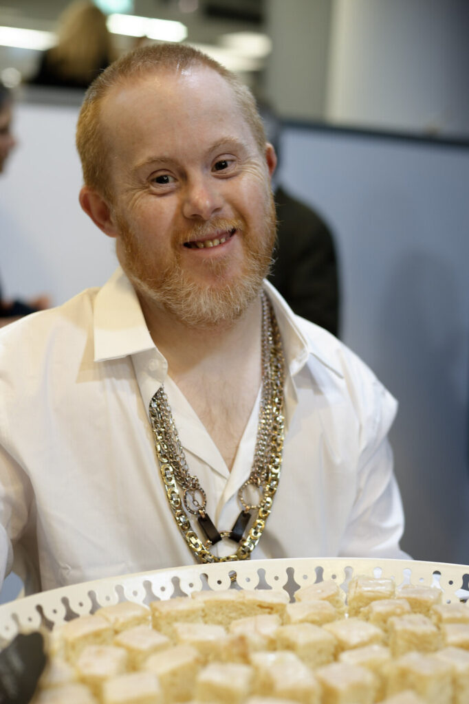Person smiling while holding a tray of small cakes at an event.