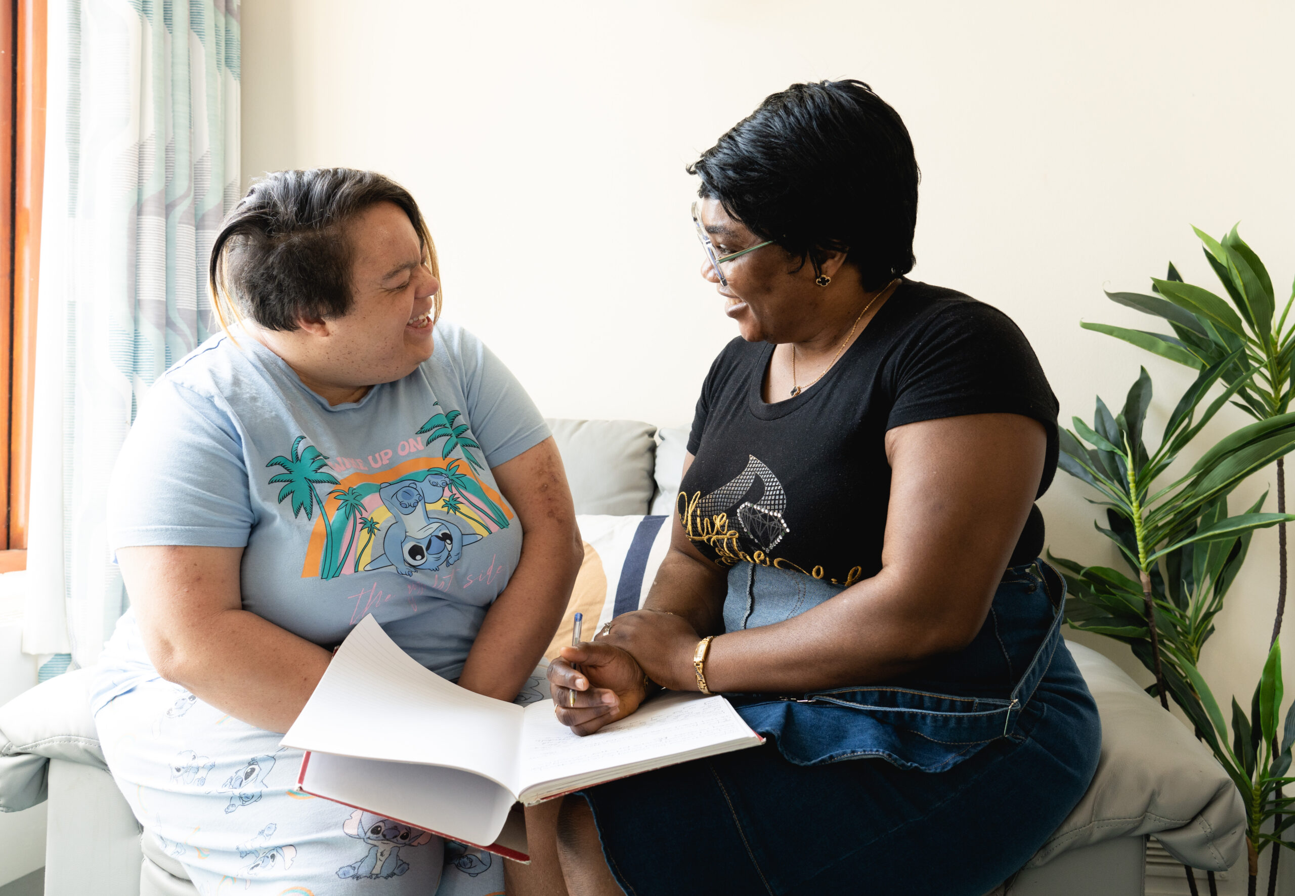 Two people sitting on a sofa smiling and looking at each other and writing care plan notes in a book