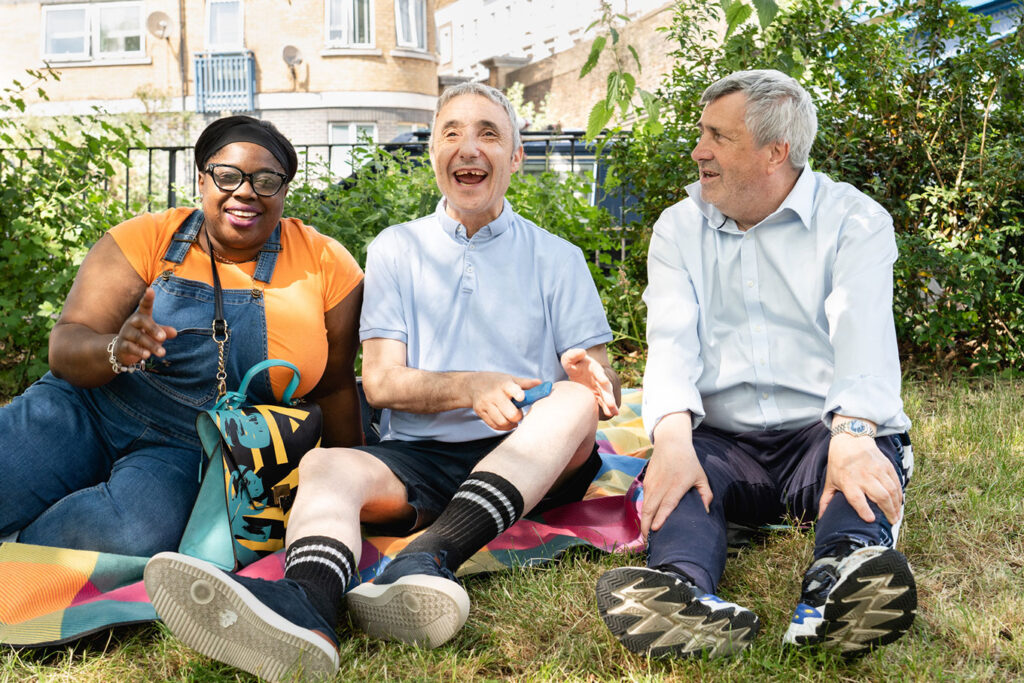 Three adults sitting on a picnic blanket in a park, laughing and chatting together.