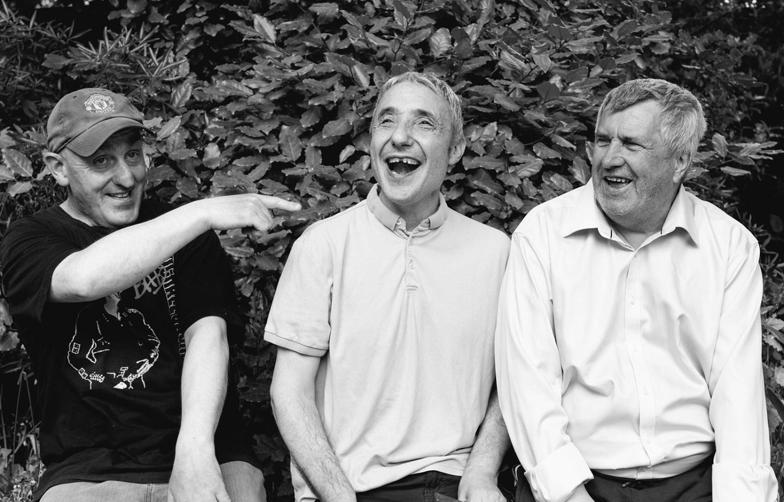 Three men are seated outside, they are laughing and one is pointing to the other