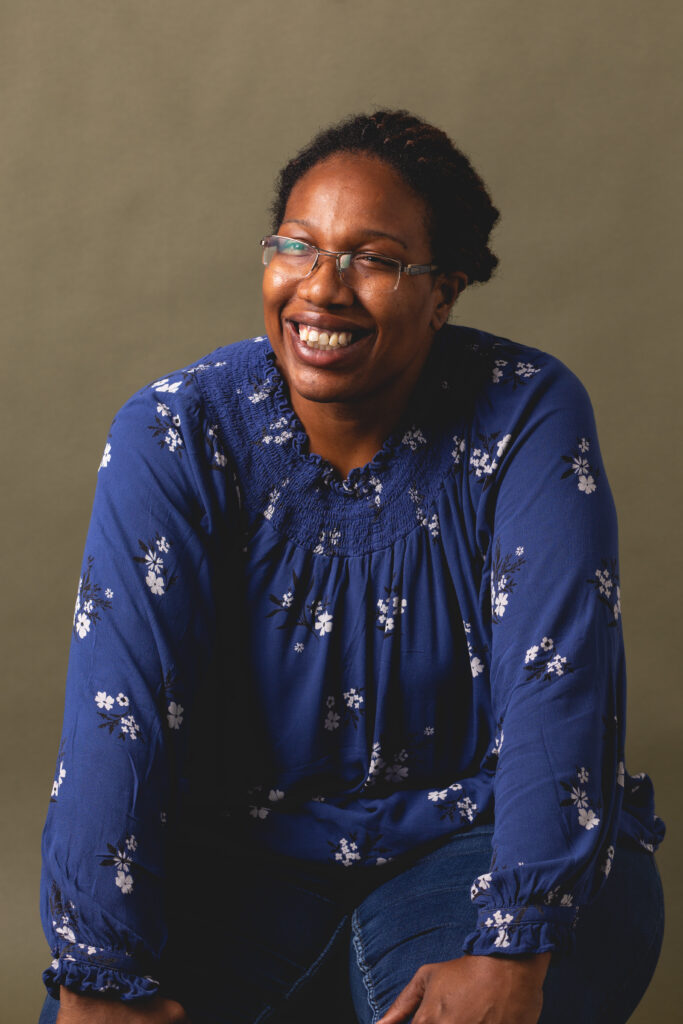 Woman wearing glasses and a blue patterned top smiling while seated in a relaxed studio portrait.