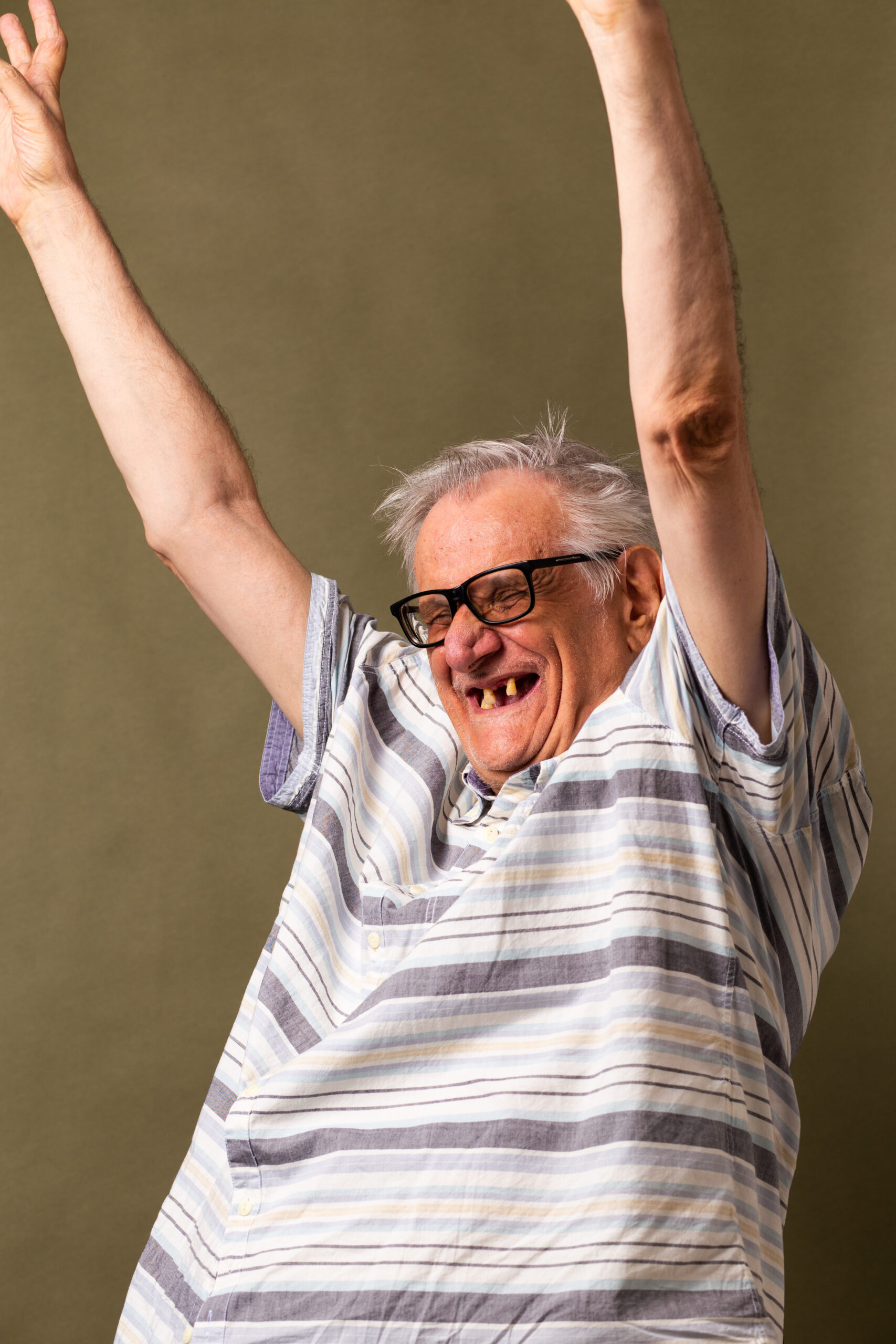 Man wearing glasses raising both arms and smiling broadly in a studio portrait.