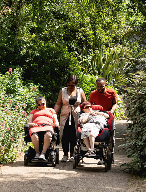 Two people in wheelchairs being pushed by support workers smileing and chatting with each other through a park with flowers and trees behind them.