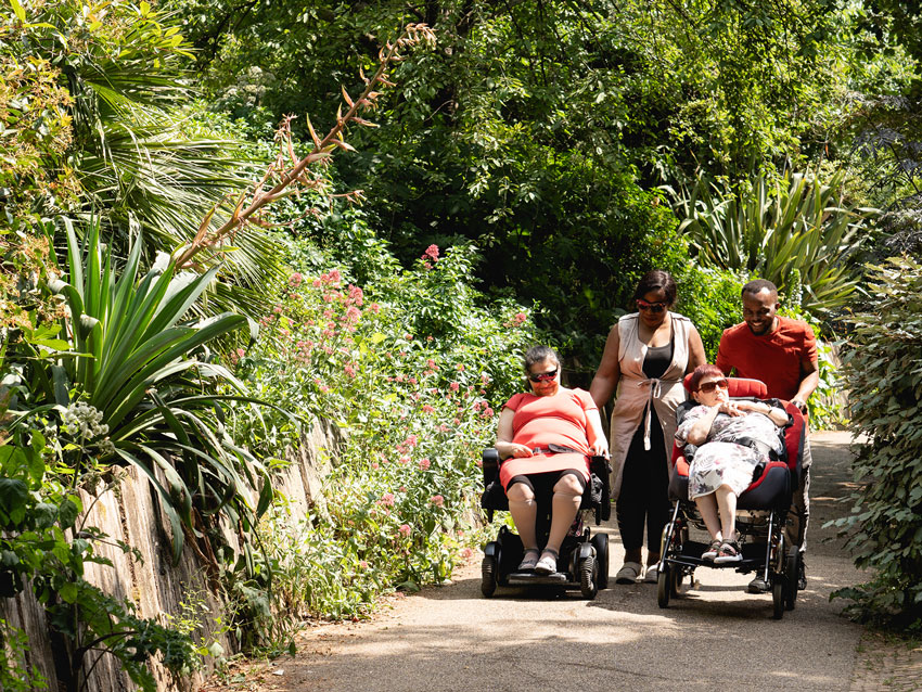 Two people in wheelchairs being pushed by support workers smileing and chatting with each other through a park with flowers and trees behind them.