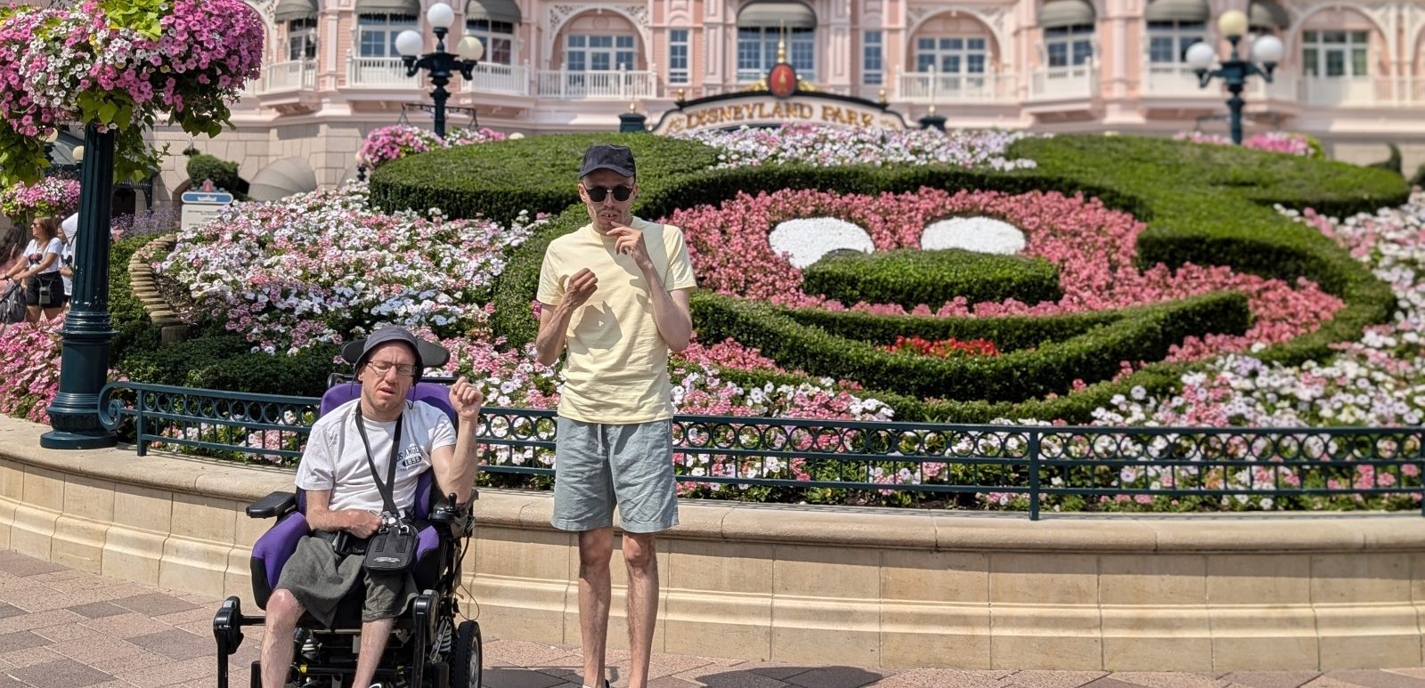 Two men in front of a Disneyland Park entrance with a flowerbed in the shape of Mickey Mouse