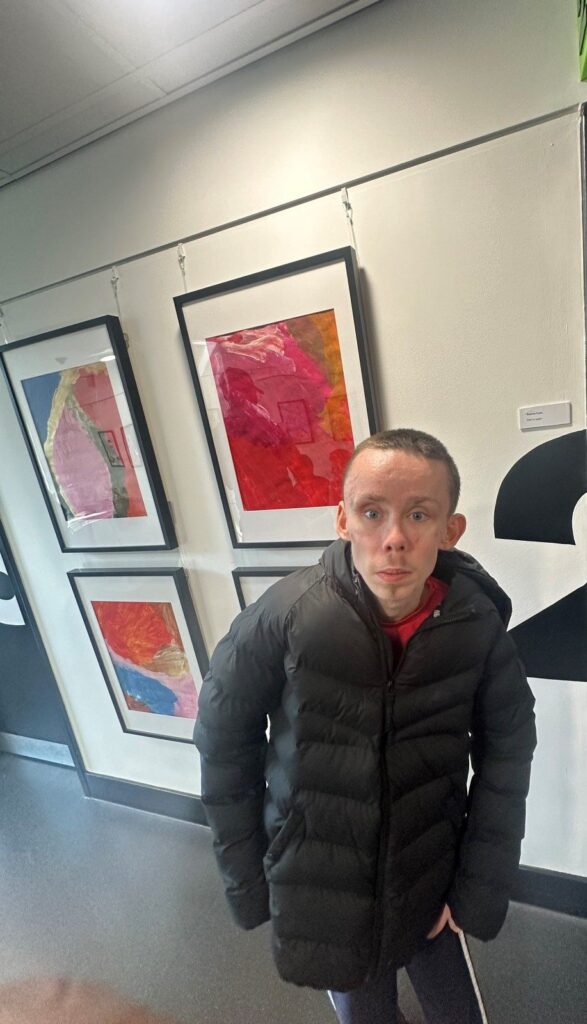 Person in a black jacket standing in front of his framed artwork on a wall.