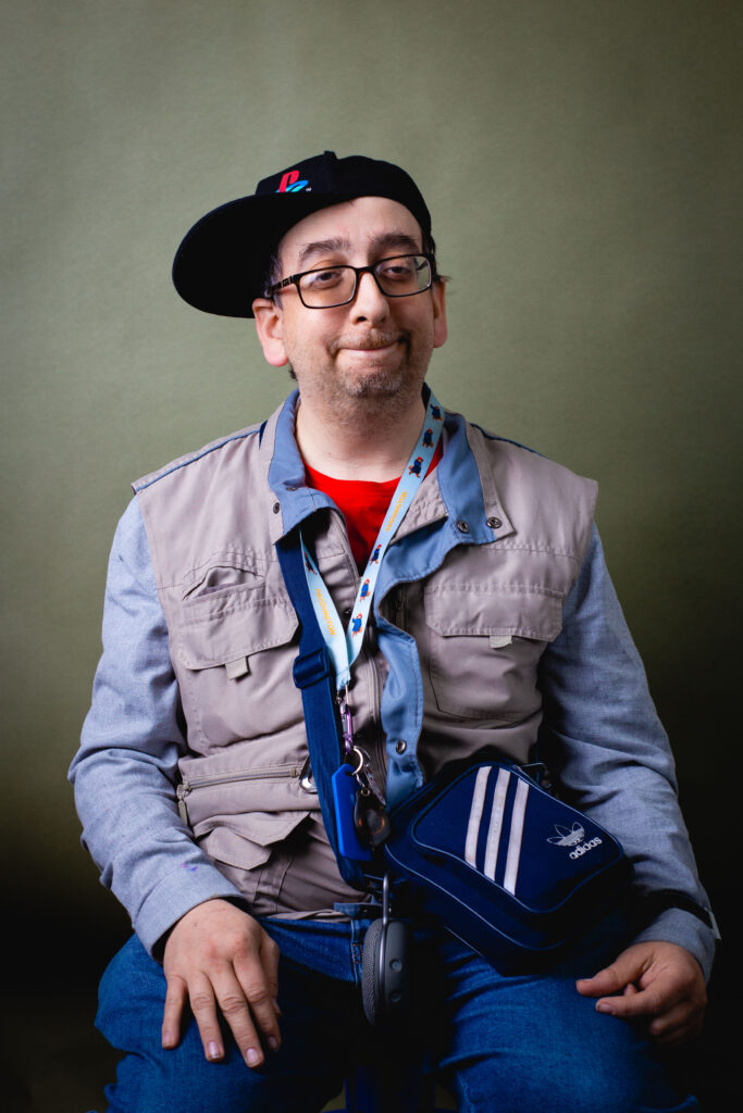 Man wearing a cap and glasses sitting for a studio portrait with a neutral expression.