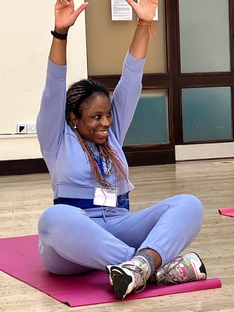Woman sitting on a yoga mat indoors with her arms raised during a group exercise session.