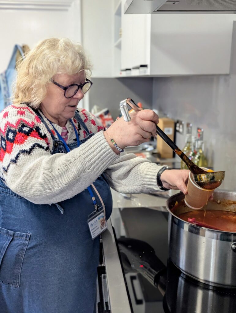 A woman serves soup with a ladle from a large pan into a paper cup