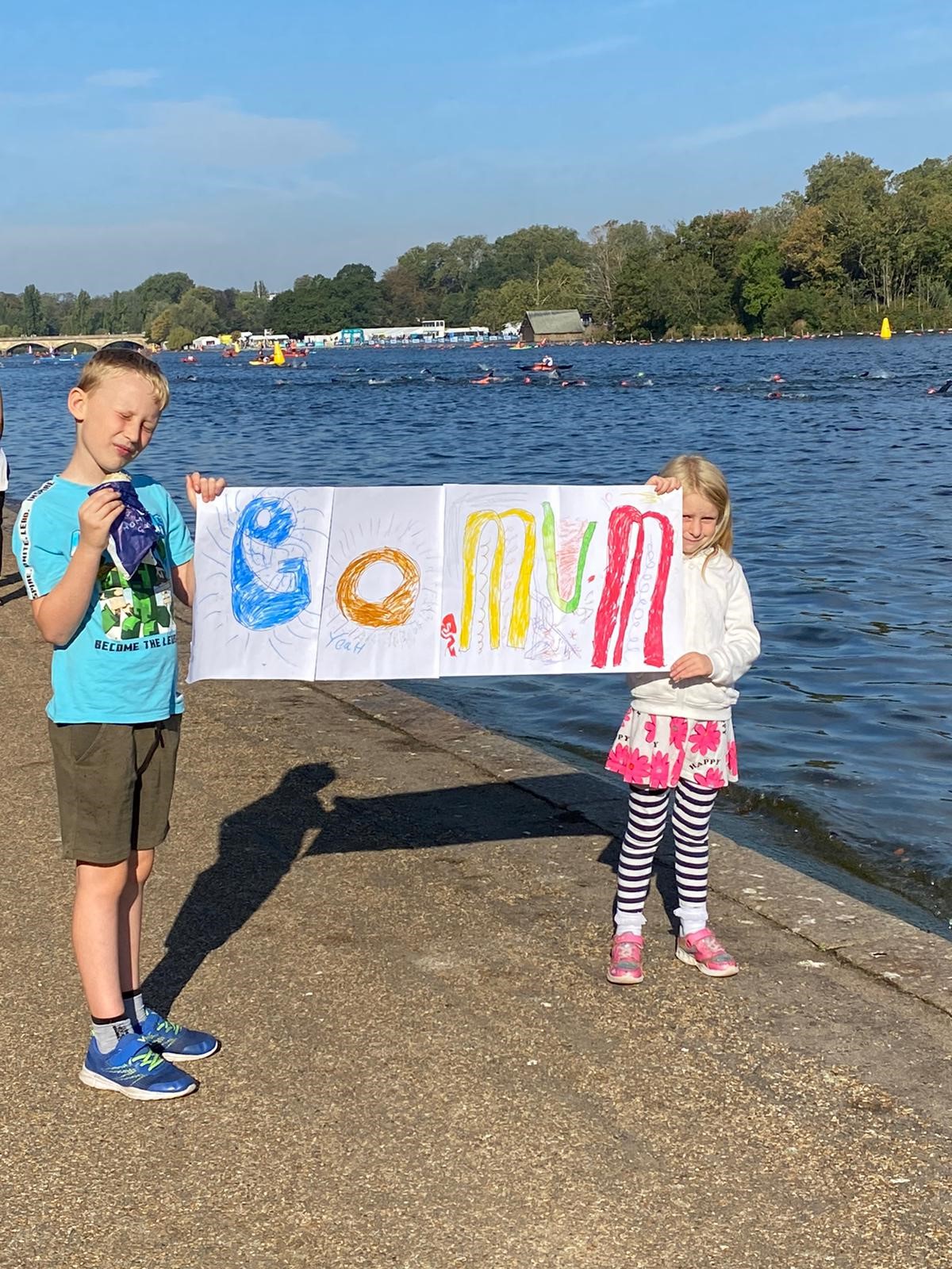 Two children standing by the water holding a handmade sign that reads 'Go Mum' to cheer on swimmers.