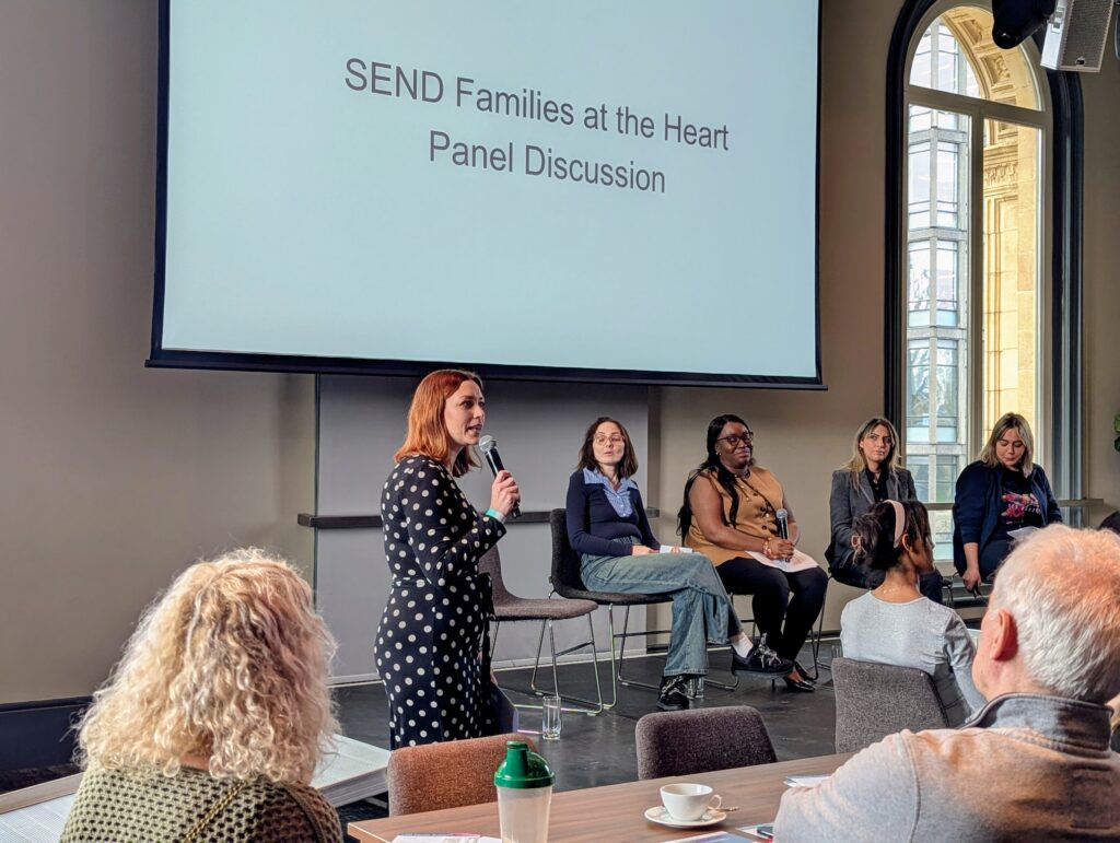 A panel, including four woman, between the ages of 35 -50 are sitting in chairs on a raised stage. A middle aged woman with gingery red hair is holding a microphone and speaking to an audience.