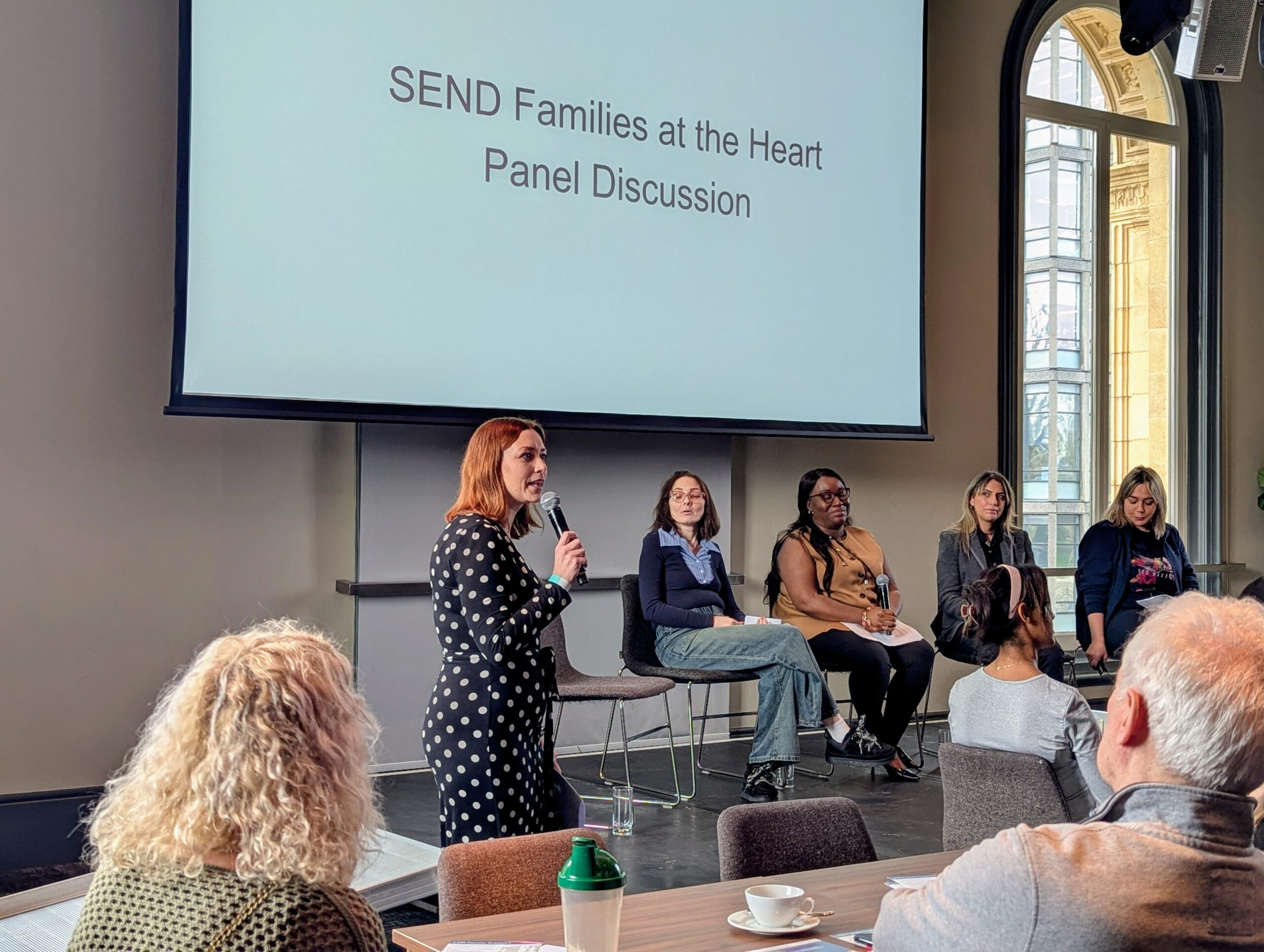 A panel, including four woman, between the ages of 35 -50 are sitting in chairs on a raised stage. A middle aged woman with gingery red hair is holding a microphone and speaking to an audience.