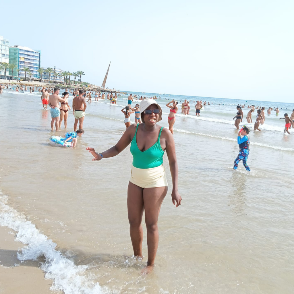 Valmie is wading in the water at the beach, as other people behind her are standing in the water. You can see lots of people at the beach, the water and an apartment. Valmie wears a white sun hat and a green top.
