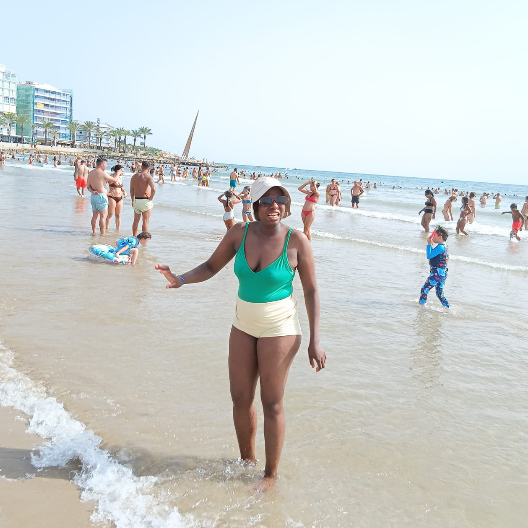 Valmie is wading in the water at the beach, as other people behind her are standing in the water. You can see lots of people at the beach, the water and an apartment. Valmie wears a white sun hat and a green top.